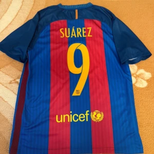 FC Barcelona Suárez 9 matchtröja Nike - Säljer en officiell FC Barcelona matchtröja från Nike med Suárez och nummer 9 på ryggen. Tröjan har klassiska blå och röda vertikala ränder, klubbmärke på bröstet och Qatar Airways som sponsor. Tillverkad i lätt och ventilerande material, perfekt för fotboll eller supporterliv.