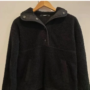 Svart fleece hoodie från Bikbok XS - Mysig svart hoodie från Bikbok i fleece med lång ärm och huva. Tröjan har en half zip med tryckknappar och två fickor framtill. Perfekt för kyliga dagar när du vill vara både bekväm och snygg.