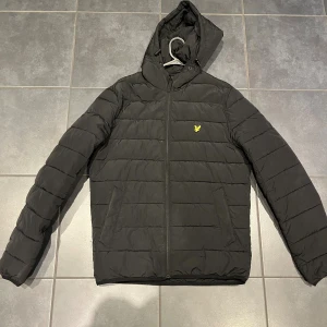 Svart pufferjacka från Lyle & Scott - Svart pufferjacka från Lyle & Scott med huva och gul logotyp på bröstet. Jackan har horisontella quiltade sömmar, dragkedja framtill och två sidofickor. Perfekt för kyliga dagar och har en clean, sportig vibe.