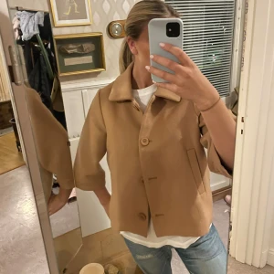 Beige ullkappa  - Kort beige kappa i ull med trekvartsärmar, klassisk krage och knäppning med stora knappar framtill. Snygg minimalistisk look med raka fickor och boxig passform. Perfekt för dig som gillar stilrena vibes.