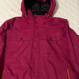 Mörkrosa vindjacka  - Snygg mörkrosa vindjacka från Everest med huva, orange detaljer vid dragkedjorna och två stora bröstfickor med knappar. Jackan har svart innerfoder och justerbar huva med snörning. Perfekt för dig som gillar färg och funktion.