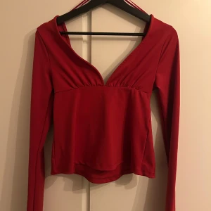 Röd långärmad omlottopp med knyt - Säljer en snygg röd omlottopp med lång ärm och djup v-ringning. Toppen har ett brett band som knyts baktill för extra stilpoäng. Materialet är mjukt och stretchigt, perfekt för att skapa en cool och trendig look. Passformen är normal och toppen är lätt croppad.