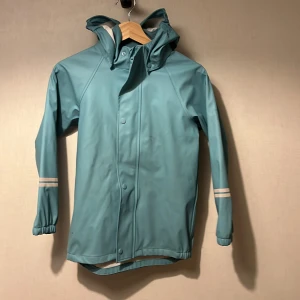 Turkos regnjacka från Kaxs - Turkos regnjacka från Kaxs med huva och reflexdetaljer på ärmarna samt en stor K på ryggen. Jackan har tryckknappar framtill och elastiska muddar vid ärmsluten. Perfekt för blöta dagar och håller dig torr oavsett väder.