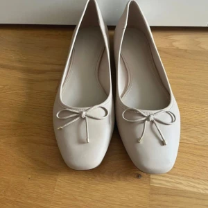 Beige ballerinaskor med rosett - Stilrena beige ballerinaskor med fyrkantig tå och söt rosettdetalj framtill. Skorna har en platt sula och är tillverkade i ett mjukt syntetmaterial som ger en klassisk och clean look. Perfekta för dig som gillar minimalistisk stil.