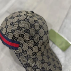 Gucci keps - Snygg keps från Gucci med klassiskt GG-mönster i beige och svart. Kepsen har en röd och marinblå rand på sidan och justerbar rem baktill. Tillverkad i bomull med insida i mörkblått tyg. Perfekt för dig som vill ha en lyxig streetstyle-accessoar.