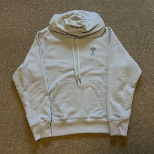 Ljusblå hoodie från AMI Paris - Säljer en stilren ljusblå hoodie från AMI Paris med broderad logga med hjärta och A på bröstet samt AMI-broderi i nacken. Tröjan har huva med snörning, känguruficka och är tillverkad i mjuk bomull. Perfekt för en chill och trendig look.