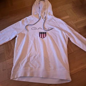 Vit hoodie från GANT med logga - Vit hoodie från GANT med stor logga och sköld på bröstet. Använd endast ett fåtal gånger.
