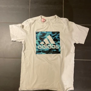 Vit Adidas t-shirt med blått tryck - Snygg vit t-shirt från Adidas med stort blått och svart grafiskt tryck på bröstet och klassisk Adidas-logga. T-shirten har rund hals och korta ärmar, perfekt för en sportig och avslappnad stil.