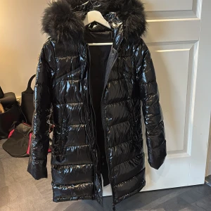 Svart glansig dunjacka med fuskpäls - Säljer en svart, glansig dunjacka med stor fluffig fuskpäls på huvan. Jackan är lång och har dragkedja framtill. Materialet är nylon och polyester, med fyllning av vitt dun. Perfekt för kalla dagar! Jackan är använd endast en vinter och är i bra skick, den är i storlek S men funkar jätte bra om man har storlek Xs. Pris går att diskuteras vid snabb affär. 