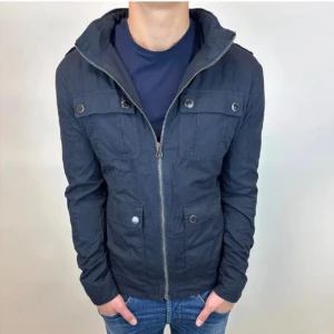 Field jacka från selected homme  - Snygg mörkblå vindjacka med huva och dragkedja framtill. Jackan har fyra fickor med knappstängning och en stilren, sportig look. Perfekt för dig som gillar enkel och clean stil. Passar till jeans och t-shirt för en chill vibe.