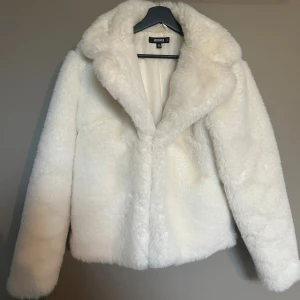 Vit pälsjacka från Missguided - Säljer en fluffig vit pälsjacka från Missguided med bred krage och långärmad design. Jackan har en mjuk och lyxig känsla, perfekt för dig som vill sticka ut med en clean och trendig look. Passformen är normal och materialet är syntetpäls.