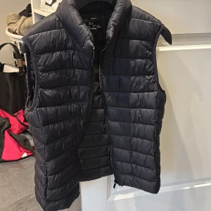 Svart dunväst XS  - Svart dunväst från primark i  storlek XS. Västen har quiltad design med horisontella sömmar, hög krage och dragkedja framtill. Perfekt för lager-på-lager och enkel att matcha med olika outfits. Västen är helt oanvänd 