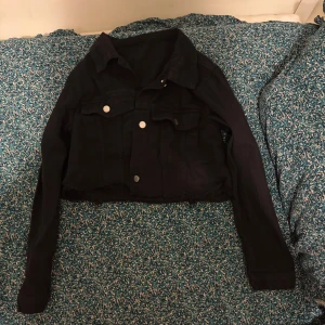 Svart croppad jeansjacka Petite M - Svart croppad jeansjacka med klassisk krage och silverfärgade knappar framtill. Jackan har två bröstfickor med lock och långa ärmar. Rå kant nedtill för en edgy vibe. Perfekt för dig som gillar streetstyle och vill ha en snygg layering-piece.