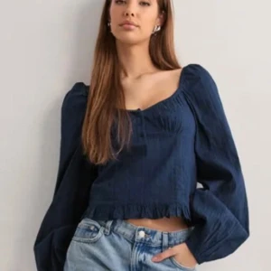 Mörkblå blus med volangkant - Supersöt mörkblå blus med långa puffärmar, v-ringad halsringning och tre knappar framtill. Blusen har en volangkant nertill och är lätt croppad. Tillverkad i ett tunt, vävt material som känns luftigt och skönt. Endast använd en gång!