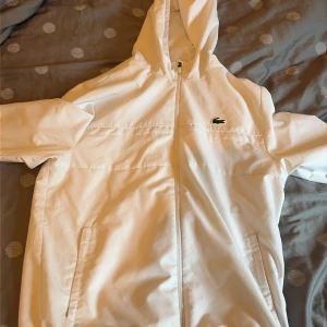 Vit vindjacka från Lacoste Sport - Snygg vit vindjacka från Lacoste Sport med huva och dragkedja framtill. Jackan har två sidofickor och det klassiska Lacoste-krokodilmärket på bröstet. Regular fit och tillverkad i lätt polyester med meshfoder för extra komfort.