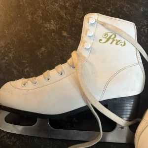 Vita konståkningsskridskor Pros 35-36 - Snygga vita konståkningsskridskor från Pros i storlek 36. Klassisk modell med snörning, högt skaft och robust klack. Ovandel i syntetmaterial och slitstarka stålblad. Perfekta för dig som vill glida fram på isen med stil.