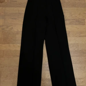 Svarta kostymbyxor från Madlady - Svarta kostymbyxor från Madlady med hög midja och raka ben. Byxorna är tillverkade i en mjuk mix av polyester, viskos och elastan som ger en skön passform. Perfekta för en clean och stilren look.