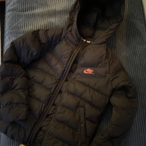 Svart dunjacka/pufferjacka från Nike strl 128-137.  - Svart dunjacka från Nike med huva och dragkedja framtill. Jackan har ett tryckt Nike-logotyp i rosa på bröstet. Perfekt för kalla dagar och har en schysst, sportig vibe.