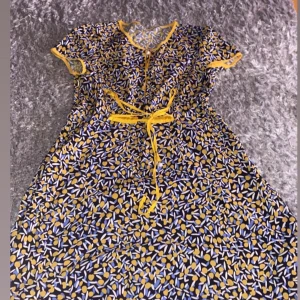 Blommig kortklänning med knytband - Säljer en söt kortklänning med blommigt mönster i gult, blått och vitt på mörk botten. Klänningen har korta ärmar, rund hals och ett gult knytband i midjan. Materialet känns lätt och luftigt, perfekt för varma dagar. Fina gula detaljer vid ärmslut och halsringning.