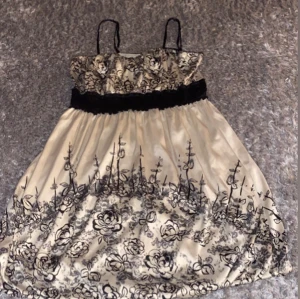 Vit bejg och svart blommig klänning Vila - Supersöt kortklänning från Vila i glansigt material med tunna axelband och svart midjeband. Klänningen är vit med svart blommigt mönster och har en lätt utställd form. Perfekt för dig som gillar romantiska detaljer och vill sticka ut med en unik look.