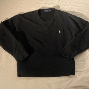 Svart sweatshirt från Polo Ralph Lauren - Svart sweatshirt från Polo Ralph Lauren med klassisk vit broderad logga på bröstet. Tröjan har rund hals, ribbade muddar och är tillverkad i mjuk bomull. Perfekt för dig som gillar stilrena och tidlösa plagg.