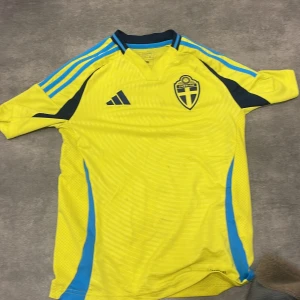 Svenska landslagets gul-blå Adidas tröja - Gul fotbollströja från Adidas med blå detaljer och Svenska Fotbollförbundets emblem på bröstet. Tröjan har korta ärmar, blå ränder på axlarna och texten 'Sverige' på ryggen. Tillverkad i lätt och ventilerande polyester, perfekt för match eller träning. Jättesnygg tröja som man borde köpa.