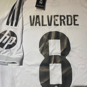 Real Madrid Valverde #8 fotbollströja M - Snygg vit Real Madrid fotbollströja från Adidas med Valverde och nummer 8 på ryggen. Tröjan har svarta detaljer, gula sömmar och klubbmärke broderat på bröstet. Tillverkad i lätt polyester med Aeroready-teknologi och korta ärmar. Perfekt för dig som älskar fotboll och Real Madrid.
