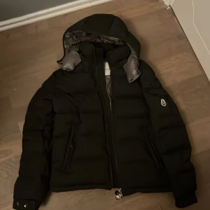 Grå Moncler pufferjacka med huva - Grå pufferjacka från Moncler med ikonisk logotyp på ärmen, huva och två dragkedjeförsedda fickor. Insidan är grå med ett coolt serietryck och märkestaggar. Jackan har en clean look och passar perfekt när du vill hålla dig varm och se fresh ut.