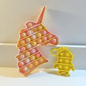 Pop It Fidget Toy Unicorn & Rocket Set - Färgglad Pop It fidget-leksak i enhörningsform samt en mindre gul raketformad Pop It med nyckelring. Perfekt för stresslindring och lek. Båda i mycket fint skick utan synligt slitage.