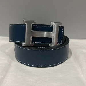 Marinblå bälte - Snyggt marinblått bälte i skinn med en stor, silverfärgad H-spänne. Bältet har vit kontrastsöm längs kanterna och en klassisk, stilren design som verkligen sticker ut. Perfekt accessoar för att lyfta din outfit med lite lyxig vibe.