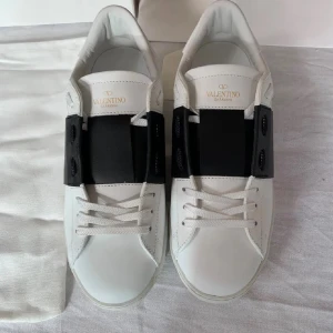 Valentino Garavani Open sneakers vit/svart - Snygga Valentino Garavani Open sneakers i vitt skinn med bred svart läderdetalj över vristen. Klassisk rund tå, vita snören och diskreta nitar baktill på sulan. Perfekta för dig som gillar stilrena och exklusiva sneakers med en twist.