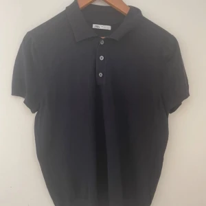 Svart pikétröja från Zara - Klassisk svart pikétröja från Zara med korta ärmar och tre knappar framtill. Tröjan har en mjuk krage och är tillverkad i bomull, perfekt för en clean och stilren look. 