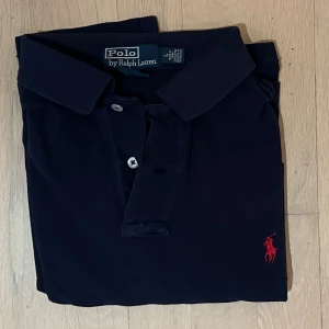 Mörkblå pikétröja från Polo Ralph Lauren - Snygg mörkblå pikétröja från Polo Ralph Lauren med klassisk krage och två knappar framtill. Tröjan har korta ärmar och den ikoniska röda Polo-loggan broderad på bröstet. Perfekt för dig som gillar stilrena och tidlösa plagg.