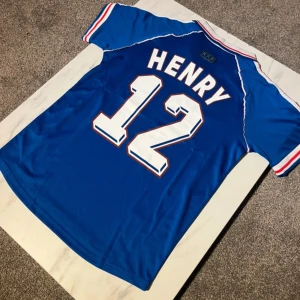 Blå Frankrike VM 98 Henry tröja Adidas - Säljer en ikonisk blå fotbollströja från Frankrikes VM 1998 med Henry och nummer 12 på ryggen. Tröjan har vita och röda detaljer, klassisk krage och korta ärmar. Materialet är lätt polyester med tryckta ränder och broderade emblem. Perfekt för dig som älskar retro och fotbollshistoria.