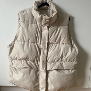 Beige dunväst från Dobber - Stilren beige dunväst från Dobber med hög krage och dragkedja framtill. Västen har två stora fickor med tryckknapp och quiltad design. Perfekt för lager-på-lager och ger en clean look till din outfit.