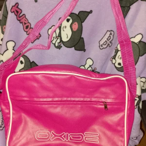 Rosa axelväska från Oxide - Cool rosa axelväska från Oxide med vit piping och dragkedja framtill. Väskan har ett stort huvudfack och justerbar axelrem. Perfekt för att bära med sig allt viktigt och ger en retro vibe till din outfit.