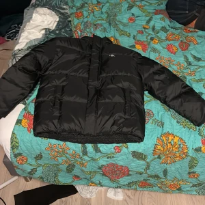 Svart pufferjacka Calvin Klein Jeans - Svart pufferjacka från Calvin Klein Jeans med logga längs ryggen och liten CK-detalj på bröstet. Jackan har lång ärm, hög krage och är tillverkad i polyester. Snygg, boxig passform och perfekt för kyliga dagar. Är helt o använd har legat i mitt rum sen förra vinter för har inte orkat lämna tillbaka den.
