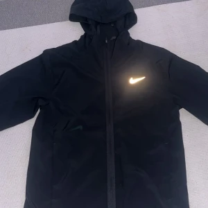 Svart Nike vindjacka med huva - Svart vindjacka från Nike med huva och reflekterande logga på bröstet. Jackan har dragkedja framtill, sidofickor med knappstängning och är tillverkad i lätt polyester. Perfekt för dig som gillar sportig stil och vill ha något funktionellt.