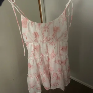 Vit och rosa blommig sommarklänning - Supersöt kort klänning i vitt med rosa blommigt mönster. Klänningen har tunna axelband med knyt, volangdetaljer och smock i ryggen för extra komfort. Perfekt för varma dagar och ger en romantisk vibe.