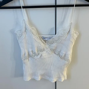 Vit ribbad linnetopp med spets Zara - Supersöt vit ribbad linnetopp från Zara med tunna axelband och vacker spetsdetalj längs urringningen och under bysten. Croppad modell med vågig nederkant, perfekt för lager på lager eller som statement-piece.