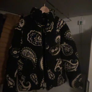Svart teddyjacka med paisleymönster FSBN XS - Säljer en svart teddyjacka från FSBN i storlek XS med fett paisleymönster i vitt och blått. Jackan har hög krage, dragkedja framtill och är riktigt mjuk och fluffig. Perfekt för dig som vill sticka ut med en unik look och hålla dig varm.