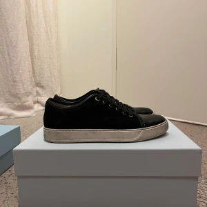 Svarta sneakers från Lanvin i mocka - Snygga svarta sneakers från Lanvin med ovandel i mocka och tå i blankt skinn. Skorna har klassisk snörning och vit platt sula med diskret logga undertill. Perfekta för dig som gillar stilrena och lyxiga detaljer.