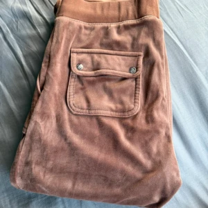 Bruna velourbyxor Juicy Couture - Mysiga bruna mjukisbyxor från Juicy Couture i velour med snörning i midjan och fickor med lock och tryckknappar bak. Byxorna har en relaxed passform och breda ben, perfekt för en chill och trendig look.