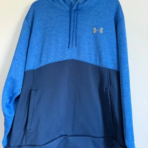 Blå hoodie från Under Armour - Snygg tvåtonad blå hoodie från Under Armour med huva och snörning. Övre delen är ljusblå med logga på bröstet, nedre delen är mörkblå. Tröjan har praktiska fickor på sidorna och en relaxed passform, perfekt för chill eller träning.