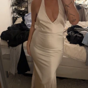 Champagnefärgad långklänning i satin - Snygg långklänning i champagnefärgat satinmaterial med djup v-ringning och öppen rygg. Klänningen har halterneck och smickrande figurnära passform. Perfekt för dig som vill sticka ut med en elegant look. Har i färgen röd, svart och champagne. 