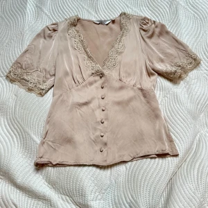 Champagnefärgad blus med spetsdetaljer - Champagnefärgad satin med v-ringning och spetsdetaljer vid halsringning och ärmslut. Blusen har korta puffärmar och knappar framtill. Märke &otherstories och storlek 34