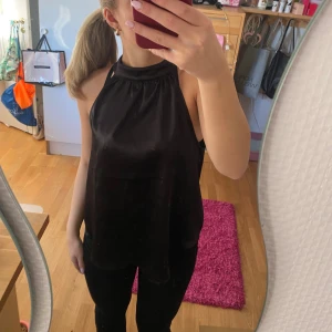Svart halterneck linnetopp  - Säljer en svart halterneck linnetopp i glansigt liknande satinmaterial. Toppen är ärmlös och har en hög hals med knäppning bak i nacken.
