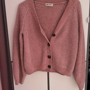 Rosa stickad kofta från H&M - Mysig rosa stickad kofta från H&M med grov ribbstickning och fyra mörka knappar framtill. Koftan har v-ringad hals och långa ärmar, perfekt för att hålla värmen under kyliga dagar. Snygg att ha både öppen och stängd.
