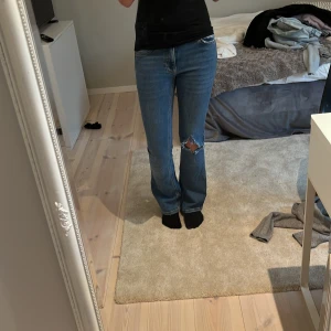 Blå bootcut jeans från Gina Tricot  - Säljer ett par blå bootcut jeans från Gina Tricot  i storlek 34. Jeansen har låg midja, klassisk femficksdesign och en snygg sliten detalj med hål på ena benet. Tillverkade i jeansmaterial med en rak och något utsvängd passform.