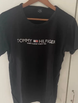 Svart t-shirt från Tommy Hilfiger  - Svart t-shirt från Tommy Hilfiger i storlek XL. Klassisk modell med rund hals och korta ärmar. Framsidan har tryckt logga och text i vitt och rött. Skön och enkel t-shirt i bomull som funkar till allt. Skriv för mer info! ABS! Pris går att diskutera.